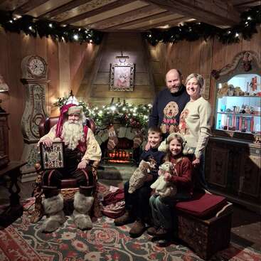 A cheerful family poses with Santa Claus in a cozy, festively decorated room. Warm lights, garlands, and a fireplace create a magical, Christmas holiday atmosphere.