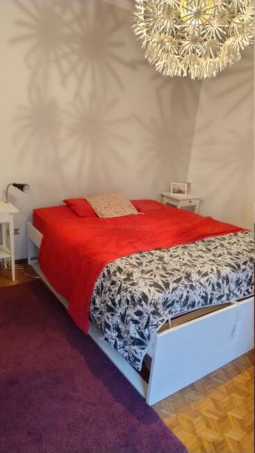 Um quarto aconchegante com uma cama coberta com roupas de cama vermelhas e em preto e branco, mesinhas de cabeceira brancas, um tapete roxo e uma luminária pendente decorativa que reproduz padrões.