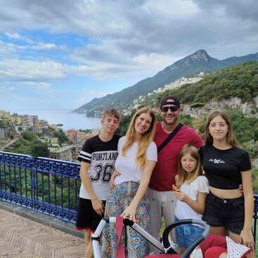 A happy family of five poses together on a scenic overlook with mountains, colorful houses, lush greenery, and the sea in the background, enjoying their vacation.