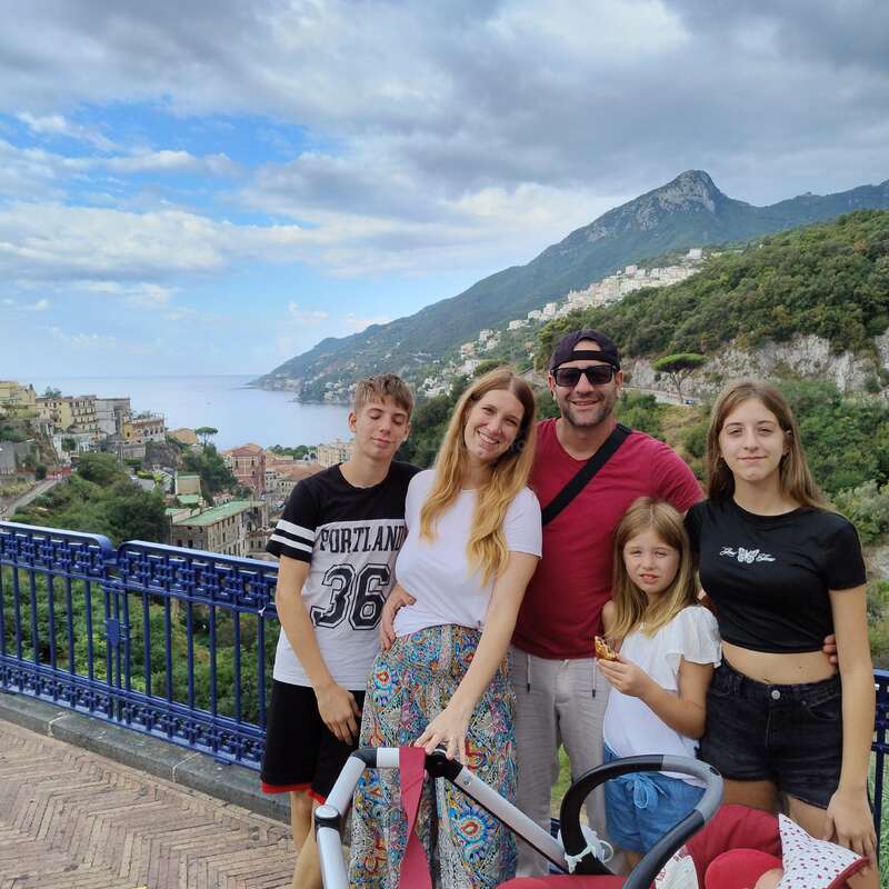 Uma família feliz de cinco pessoas posa junta em uma vista panorâmica com montanhas, casas coloridas, vegetação exuberante e o mar ao fundo, aproveitando suas férias.