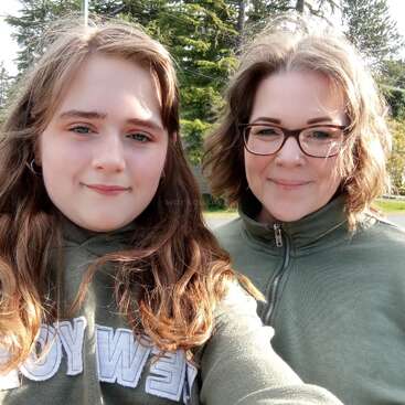 Zwei Frauen mit hellbraunen Haaren lächeln draußen. Beide tragen grüne Oberteile und stehen dicht beieinander. Hohe immergrüne Bäume und Sonnenlicht bilden einen hellen Hintergrund.