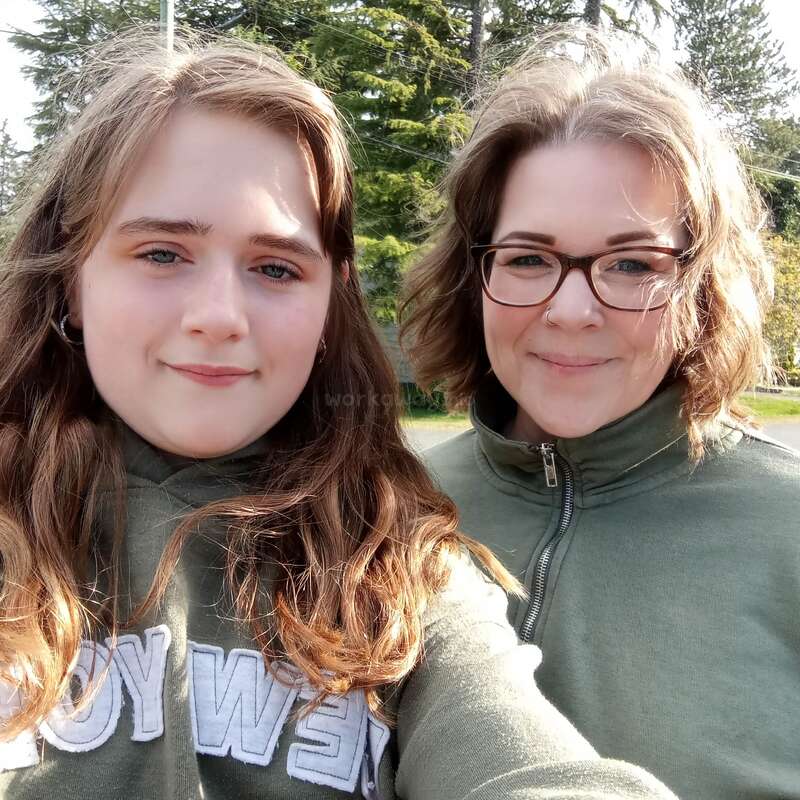 Dos mujeres de cabello castaño claro sonríen al aire libre. Ambas llevan camisetas verdes y están muy juntas. Los altos árboles de hoja perenne y la luz del sol crean un fondo luminoso.