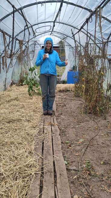 Una persona con chaqueta azul, de pie sobre tablones de madera en el interior de un invernadero, sostiene una taza y levanta el pulgar, rodeada de plantas secas y paja.