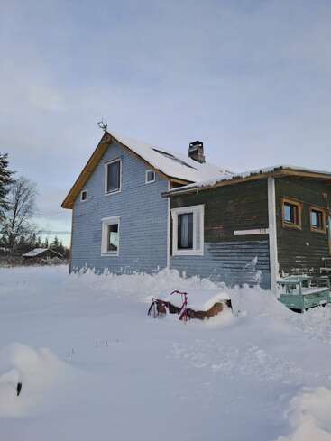 A light blue wooden house stands in deep snow, with a red bicycle partially buried. Snow blankets the yard and roof, under a cloudy winter sky.