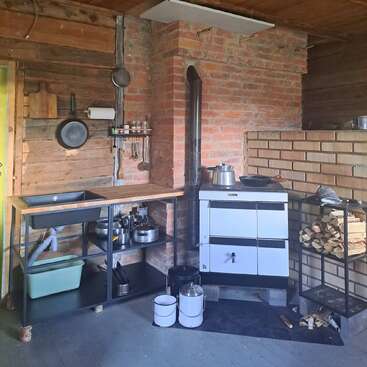 This rustic kitchen features a wood-burning stove, a small sink, stacked firewood, brick and wood walls, hanging utensils, metal cookware, and simple, practical shelving.