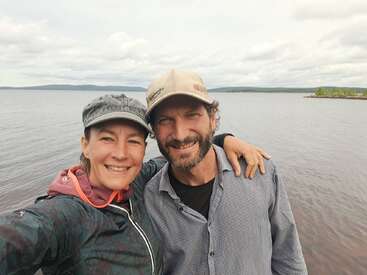 A smiling couple poses for a selfie by a calm lake under a cloudy sky. Their arms are around each other, capturing a joyful outdoor moment.
