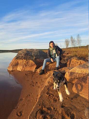 A woman sits on rocks by a calm lake, under a blue sky, while a playful black and tan dog runs energetically toward the camera on sandy ground.