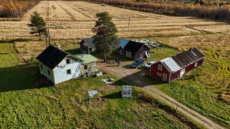 This aerial photo shows a rural homestead with four buildings, cars parked outside, surrounded by fields, trees in the distance, and a dirt road leading in.