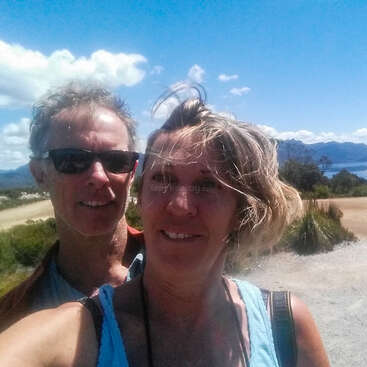 The image shows a selfie of a man and woman standing outside on a sunny day, with a scenic background of mountains and a body of water.