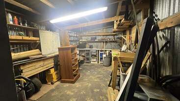 Esta imagen muestra un taller desordenado con estanterías de madera, paredes metálicas, una luz fluorescente, diversas herramientas, equipos y materiales esparcidos por todas partes, creando un espacio de trabajo funcional.