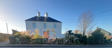 A white two-story house with two chimneys, surrounded by a stone wall, garden with tall grasses, under a clear blue sky on a sunny day.
