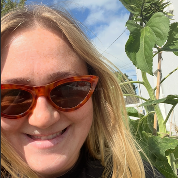 A smiling woman with blonde hair and tortoiseshell sunglasses takes a selfie outdoors beside a tall sunflower plant under a bright blue sky with clouds.