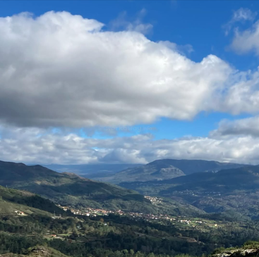 Rolling green mountains stretch into the distance under a dramatic sky filled with large, white clouds, while a small village nestles in the valley below.