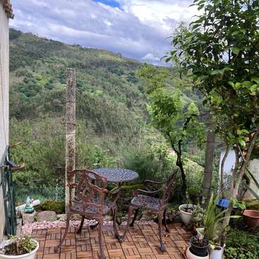 A small patio with ornate metal chairs and table overlooks a lush green mountain landscape. Potted plants surround the cozy spot under a partly cloudy sky.