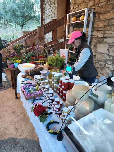 Uma mulher com um boné rosa vende geleias caseiras, mel, queijos, plantas e artesanato em uma barraca de mercado rústica ao ar livre com um muro de pedra como pano de fundo.