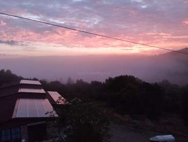Um amanhecer sereno banha o céu em tons de rosa e roxo, a névoa cobre as árvores, os painéis solares brilham e as linhas de energia se estendem pela cena tranquila.