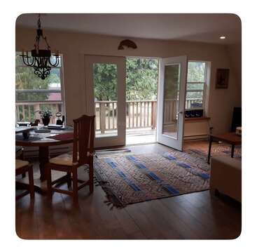 Cozy living room with wooden floors, patterned rug, round dining table, and chairs. Large windows and open doors reveal a view of trees and deck outside.