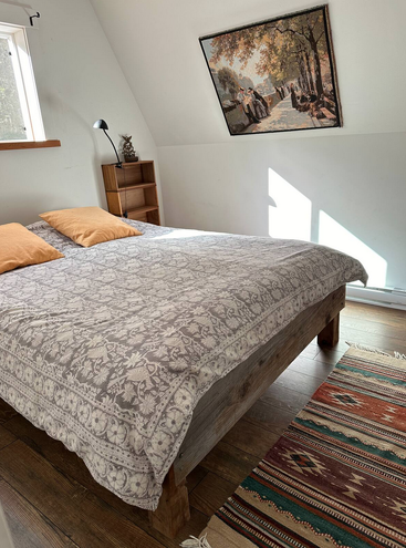 Sunlit cozy bedroom with a rustic wooden bed, two mustard pillows, patterned gray bedding, colorful woven rug, small shelf, and a painting of a park scene.
