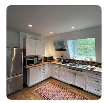 This cozy kitchen features white cabinets, wood countertops, stainless steel appliances, a colorful rug, a large window with a scenic view, and recessed ceiling lighting.