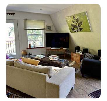 A cozy living room features a beige sofa, colorful pillows, a large TV, coffee table, armchair, artwork, wooden floors, blinds, and natural sunlight.