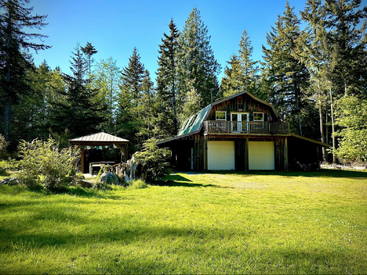 A rustic wooden house with two garage doors sits in a grassy clearing, surrounded by tall trees. A gazebo is nearby under bright, clear blue skies.
