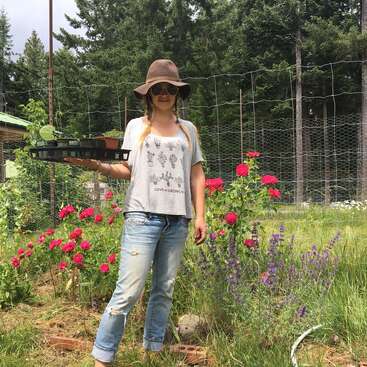 Uma mulher está em um jardim segurando uma bandeja de plantas, usando um chapéu de abas largas, óculos escuros e roupas casuais, cercada por flores e vegetação.