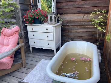A rustic outdoor bathing area features a vintage bathtub with floating flowers, a wooden chair with a pink cushion, a white dresser, potted plants, and cozy decor.