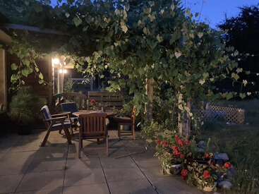 A cozy outdoor patio at dusk, featuring wooden chairs and table beneath leafy green vines. Warm light illuminates the area, with potted red flowers adding charm.