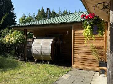 A wooden building with a green metal roof features a barrel sauna outside, surrounded by greenery, flowers in a hanging basket, and a peaceful forest background.