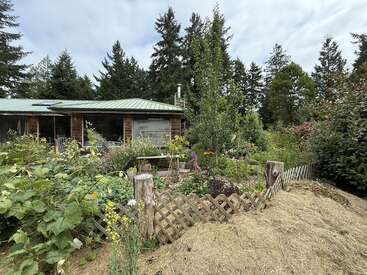 A cozy house with a green roof is surrounded by a lush, colorful garden, wooden fence, trees, and blooming flowers, creating a peaceful, rustic atmosphere.