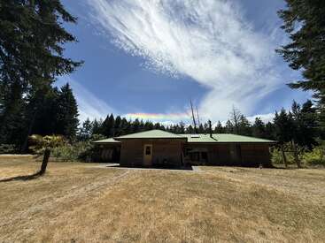 A rustic wooden cabin with a green roof sits in a clearing surrounded by trees. Above, dramatic clouds part to reveal a faint rainbow arc.
