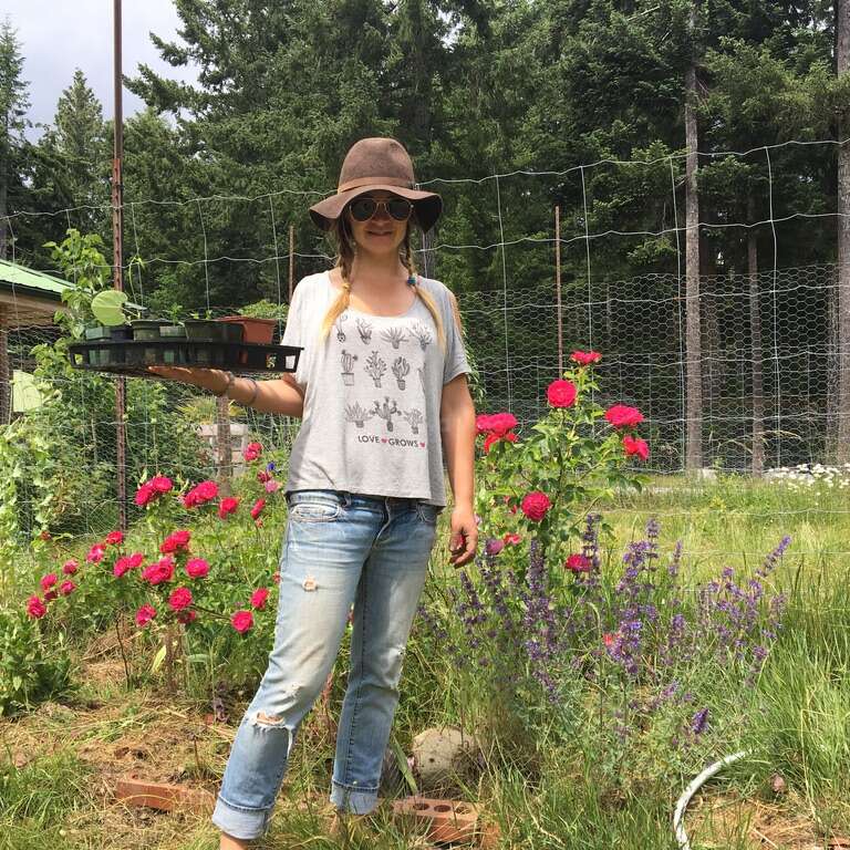 A woman stands in a garden holding a tray of plants, wearing a wide-brimmed hat, sunglasses, and casual clothes, surrounded by flowers and greenery.