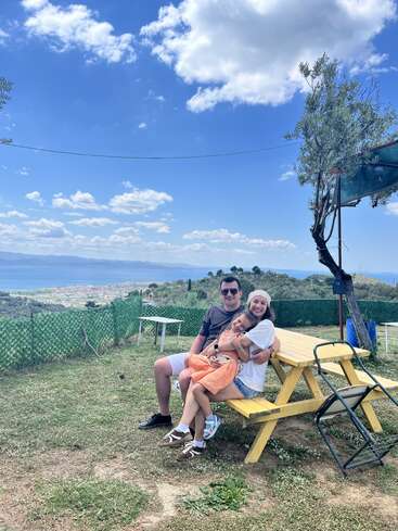 A happy family sits together on a yellow picnic bench outdoors, enjoying a sunny day with blue skies, mountains, and a distant town in the background.