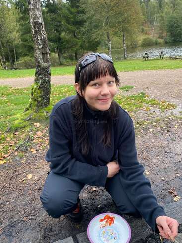 A young woman with dark hair and sunglasses on her head squats outdoors beside a plate with food, smiling, with a forest, pond, and park benches behind.