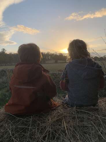 Two children, bundled in jackets, sit together on a haystack, peacefully watching a golden sunset over open fields and distant trees, embracing tranquility and friendship.