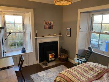 This cozy room features two large windows, a wood stove fireplace, a rocking chair, a bed with patterned blanket, a desk, and warm, neutral décor.