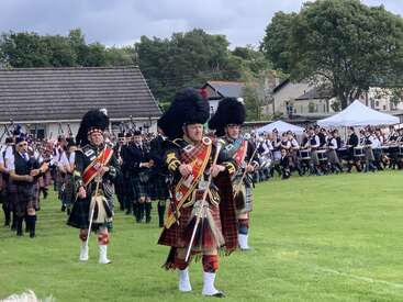 Men in traditional Scottish Highland dress lead a large parade of bagpipers and drummers, marching across a grassy field at an outdoor festival or gathering.