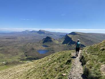 A lone hiker walks along a narrow trail on a grassy hillside, overlooking stunning mountains, valleys, and small lakes under a clear, bright blue sky.