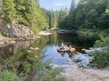 A tranquil forest river scene, sunlight filtering through trees. Two people sit on rocks in the water, surrounded by lush greenery, sandy shore, and calm reflections.