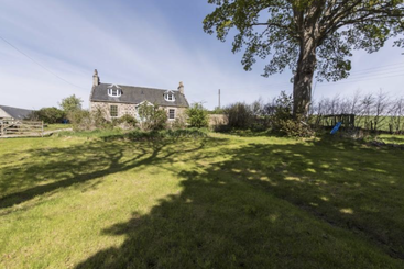 A charming stone cottage sits under a bright blue sky, surrounded by a lush green lawn, shade from a large tree, and peaceful rural scenery.