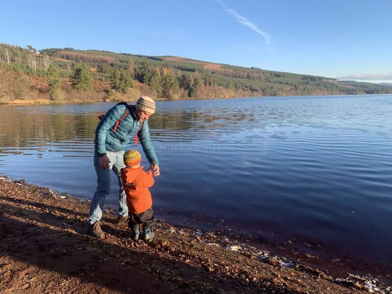 A man and child stand by a calm lakeshore, surrounded by hills and trees. Both wear jackets and hats, enjoying a peaceful, sunny outdoor moment together.