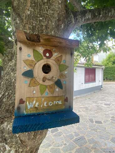 L'image représente un nichoir en bois avec un soleil et un panneau "Welcome", attaché à un tronc d'arbre devant un bâtiment blanc aux volets rouges.