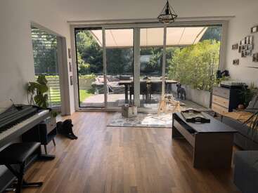 Un luminoso salón con suelo de madera, piano y grandes puertas acristaladas que dan a un soleado patio. Un perro negro está tumbado junto a la ventana.