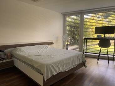 El dormitorio, acogedor y minimalista, cuenta con una cama bien hecha, un escritorio con monitor, una silla, un ventilador y grandes ventanales que permiten la entrada de luz natural y vistas a la vegetación.