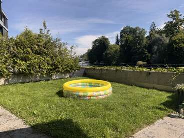 Una pequeña piscina hinchable para niños sobre la hierba verde de un soleado jardín, rodeada de arbustos y árboles, con un río y un cielo azul al fondo.