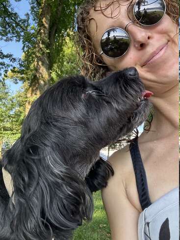 A woman wearing round sunglasses smiles as a black dog licks her cheek. They are outdoors, surrounded by trees, enjoying a playful and affectionate moment together.