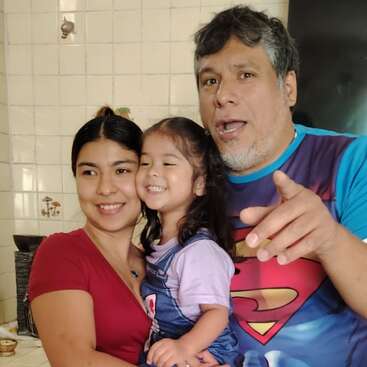 Ein fröhlicher Familienmoment: Eine lächelnde Frau hält ein glückliches kleines Mädchen im Arm, während ein Mann in einem Superman-Shirt spielerisch in die Kamera zeigt. Herzliche Atmosphäre.