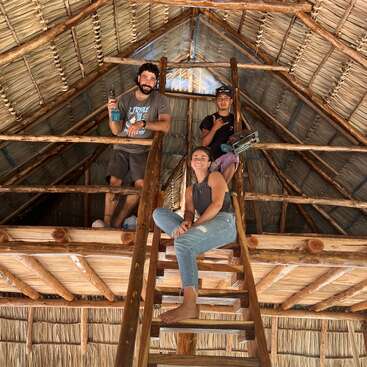 Três pessoas posam dentro de um loft rústico de madeira com telhado de palha, sorrindo e segurando ferramentas, sugerindo trabalho em equipe e construção em um ambiente tropical ou rural.