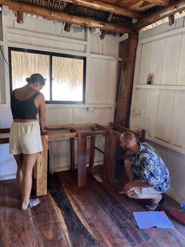 Uma mulher e um homem trabalham juntos montando móveis de madeira em uma sala rústica com piso de madeira, paredes brancas e telhado de palha, iluminada por luz natural.