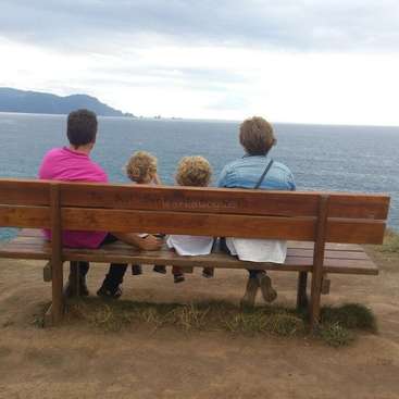 The image depicts a family of four sitting on a wooden bench overlooking the ocean, with a cloudy sky and a mountainous landscape in the distance.